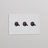 Interruptor conmutador de palanca x3 en marco doble pintable y bronce sobre pared blanca, elegante y funcional.