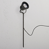 Cambridge, reflector de suelo en bronce para exteriores, con estaca y cable negro sobre pared blanca.