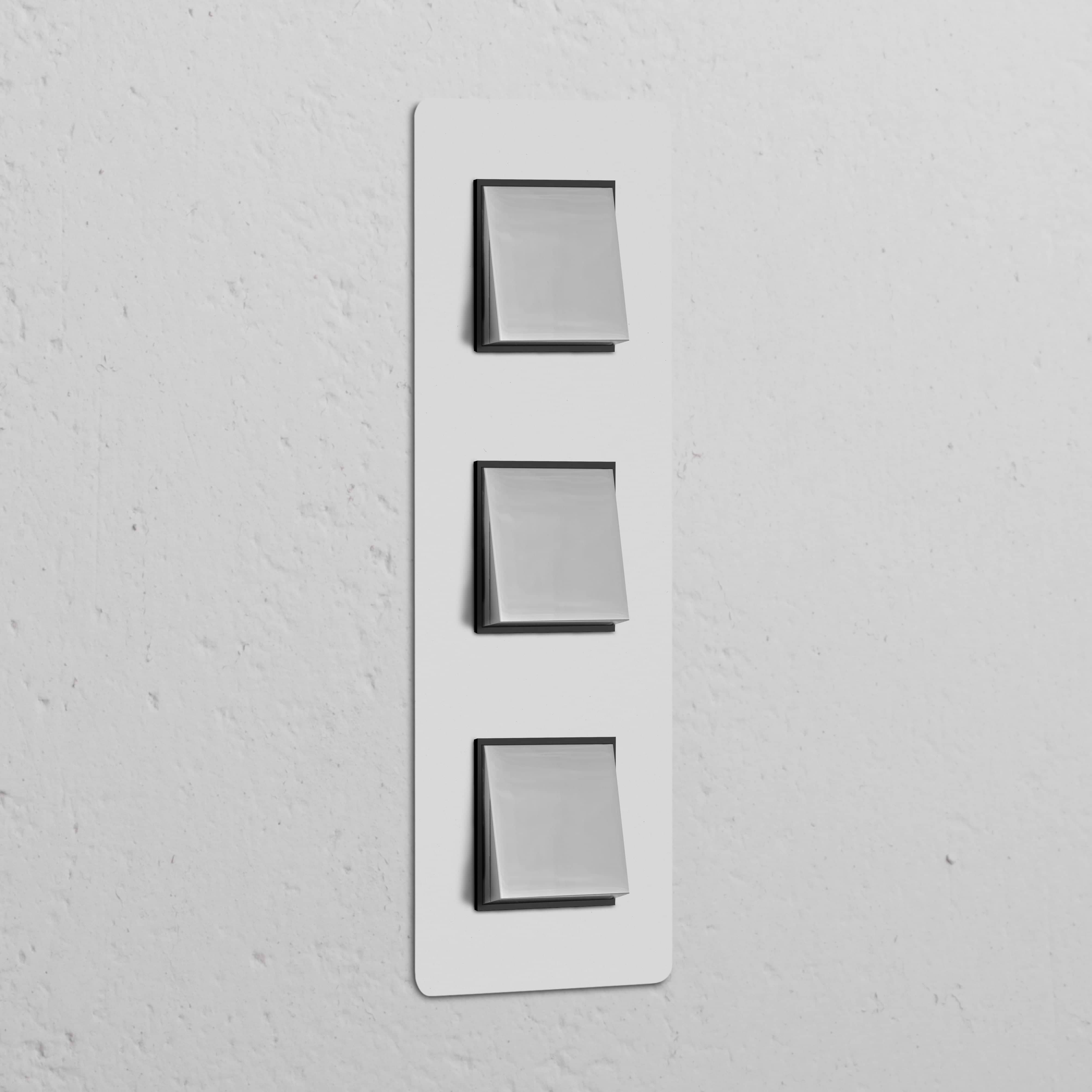 Marco interruptor basculante triple vertical - Acabado pintable y níquel pulido con inserto negro sobre pared blanca.