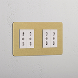 Modulo Doppio 4x Bipasso in ottone anticato e finitura bianca su una piastra beige contro una parete strutturata.