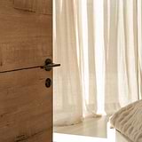 Bronze Warwick Ket Escutcheon fitted to a wooden door beneath a Bronze Door handle surrounded by cream curtains, floor and bedding. 