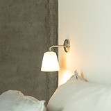 Richmond Small Wall Light in Fine Porcelain with polished nickel, mounted on a concrete wall near a bed with white pillows.
