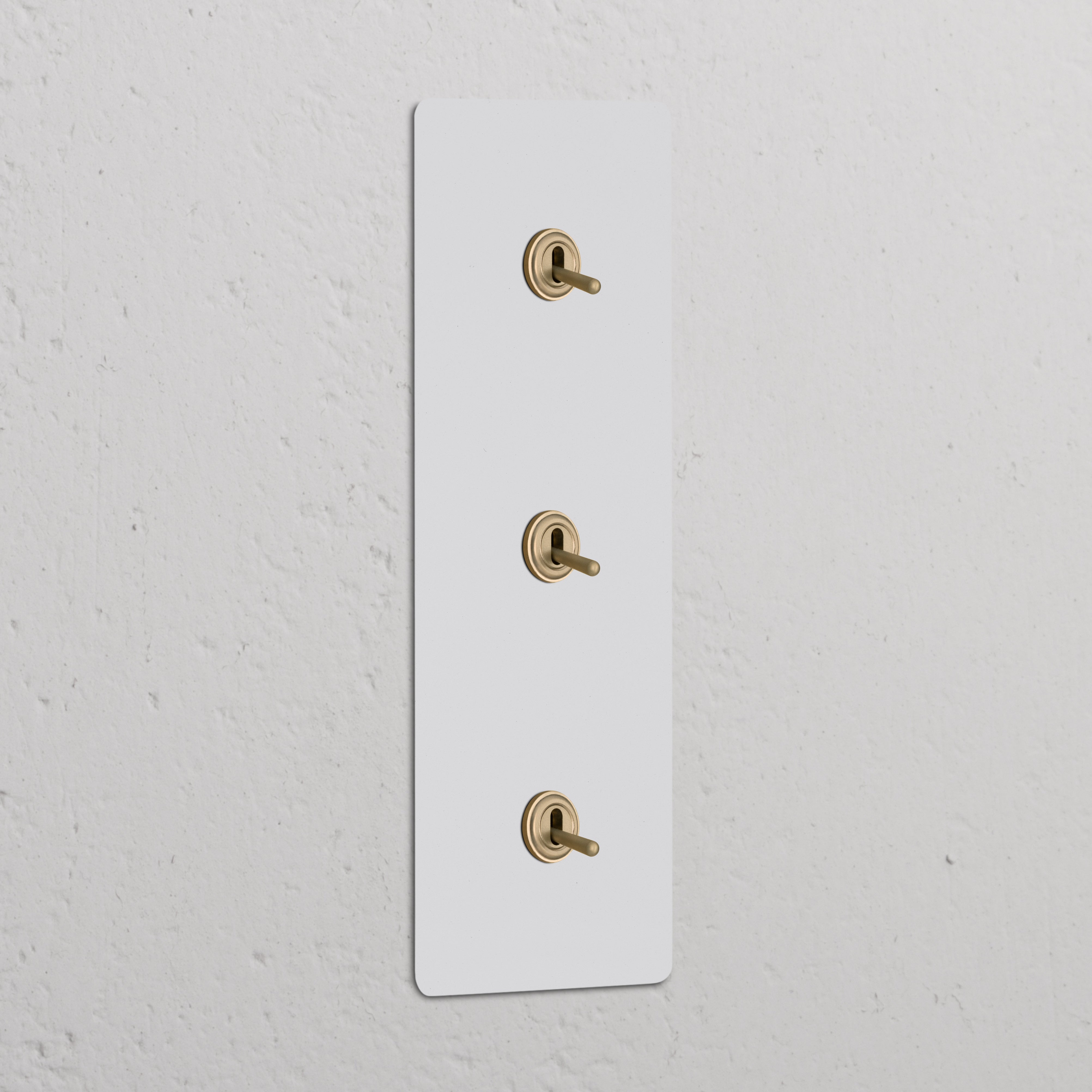 Triple 3x vertical toggle switch in paintable antique brass, mounted on a white wall plate against a textured wall.