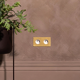 Double 2x Toggle & French Module in Antique Brass White for Double Sockets; shown on a brown wall beside a plant.
