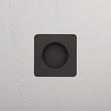 Black square wall fixture with bronze-edged circular center, mounted on a textured white wall.