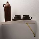 Antique Brass Brixton Shelf Bracket 150mm supporting a white marble stone shelf fixed onto a textured white wall with dark coloured pottery placed on top. 