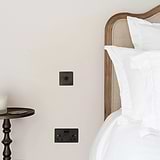 Bronze 1G two way toggle switch on white wall next to wooden framed headboard and white bedding 