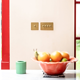 Antique brass 1G two way toggle switch fixed to cream wall next to red window frame with peach fruit bowl in the forefront 