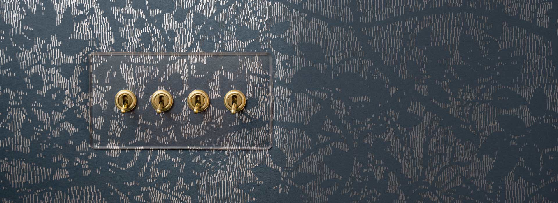 Durchsichtige Schalterplatte mit vier Kipphebeln mit der Oberfläche Antikes Messing auf einer blausilbrigen Tapete mit Blumenmuster