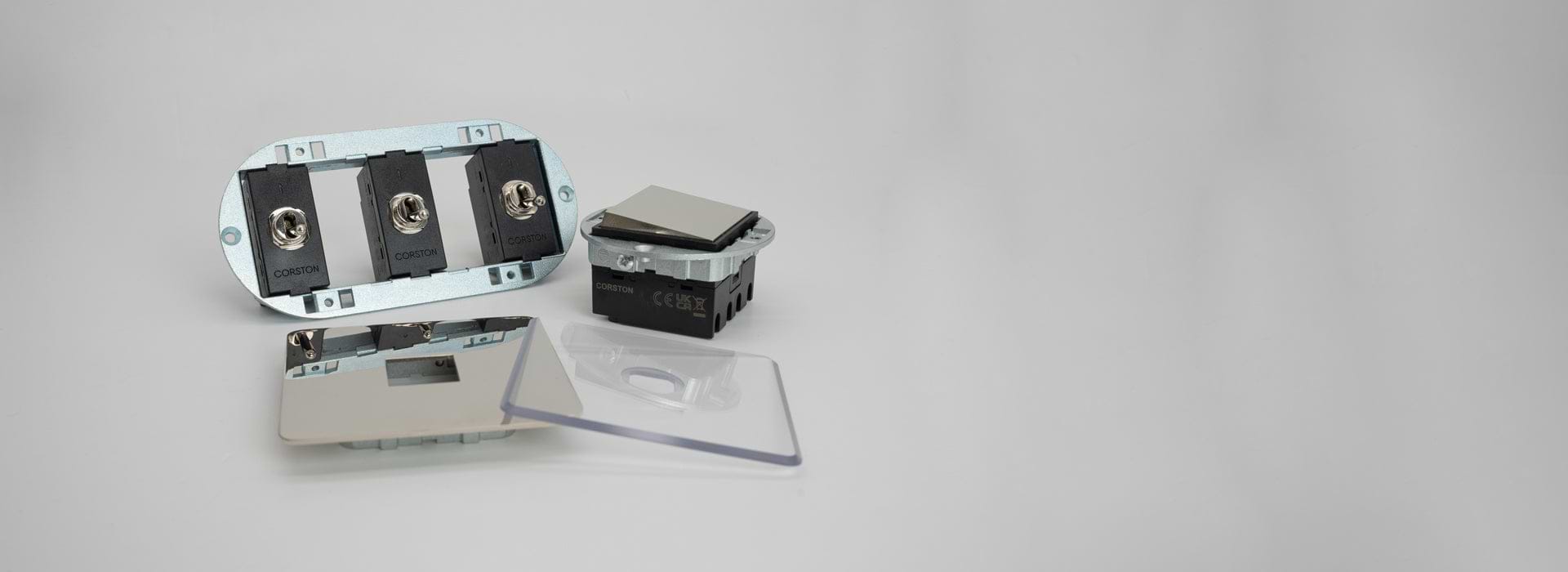 Einsatz mit drei Kipphebelschaltern, eine Wippe und eine Grundplatte aus Nickel sowie eine durchsichtige Grundplatte auf grauem Hintergrund