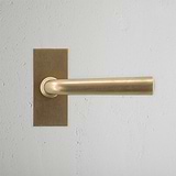 Apsley Korte Plaat Verende Deurklink - Antiek Messing op een getextureerde lichtgrijze muur, met elegant design.