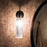 Brons Claremont kleine wandlamp met geribbeld glas, gemonteerd op een met bloemen behangen muur met een badkamermeubel naar rechts verschoven. 