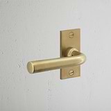 Apsley Korte Plaat Vende Deurklink - Antiek Messing op een getextureerde witte muur, elegant deurklinkontwerp.