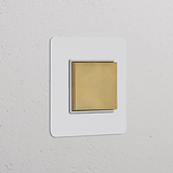 Wandschalter mit zentrierter Wippe in einem einfachen Rahmen, lackierbares Finish in Antikmessing und Weiß, auf einer strukturierten Wand.