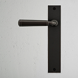 Digby gefederter Türgriff langes Schild – Bronze, mit rechteckiger Rückplatte, auf strukturierter weißer Wand.