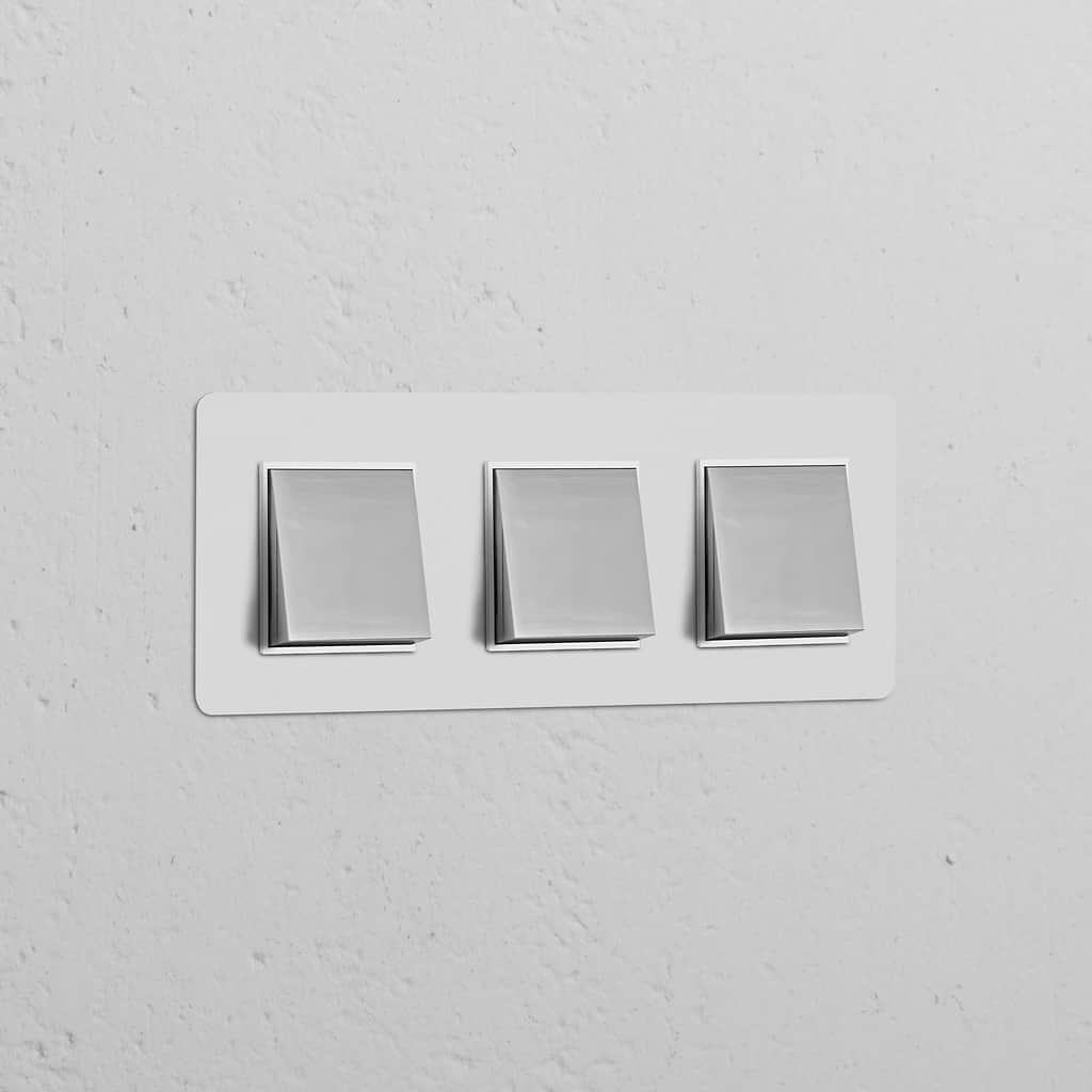 Dreifachrahmen mit 3x Wippschalter in lackierbar, poliertem Nickel und Weiß auf strukturierter weißer Wand.