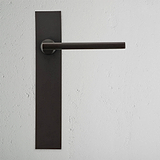 Clayton Fixer Türgriff langes Schild in Bronze auf einer rechteckigen Rückplatte vor einer weißen strukturierten Wand.