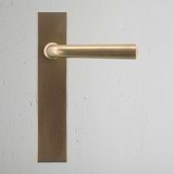 Apsley Long Plate Sprung Door Handle in antique brass finish on a textured white wall.