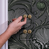 Hand operating a clear antique brass 4G vertical two-way toggle switch on a floral-patterned wall.