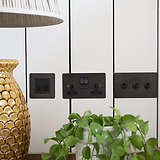 Bronze and black CAT6 socket fixed to white wooden slatted wall next to three way toggle switch with cream lampshade to the left and hanging plant in the front