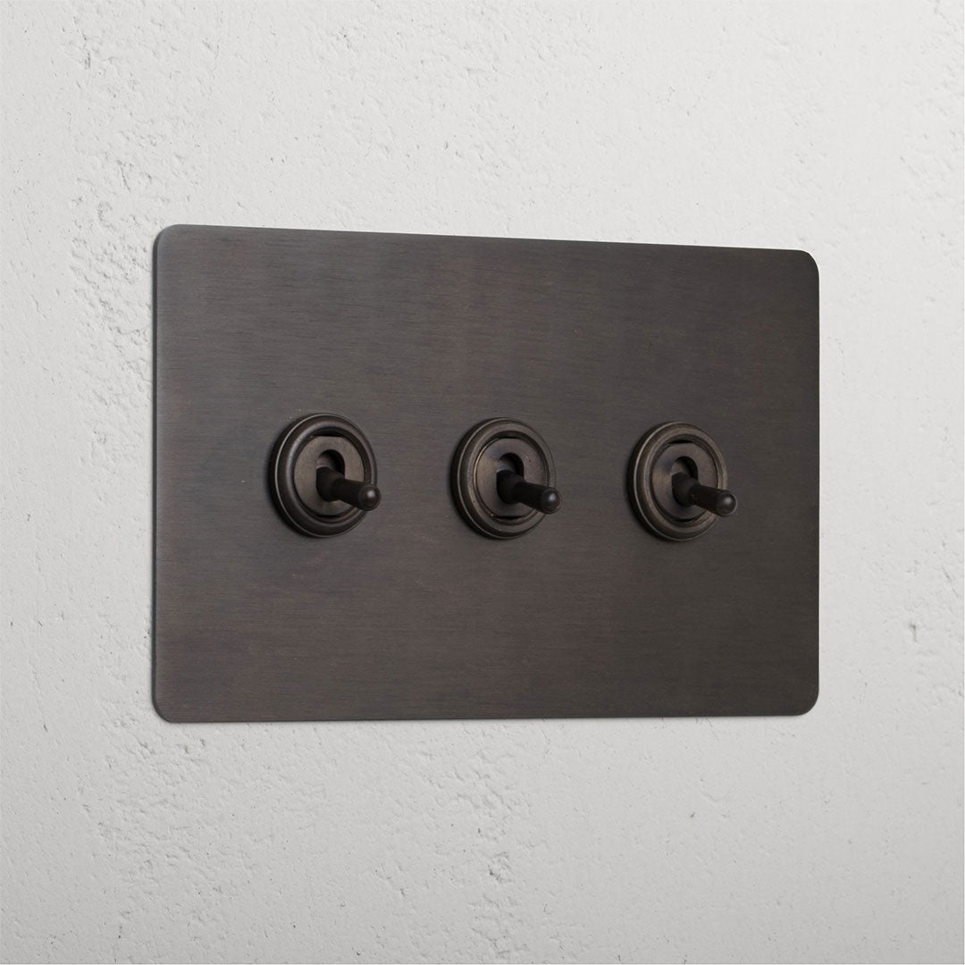 Bronze panel with three retractive toggle switches (3G) mounted on a white wall.