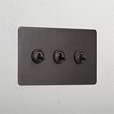 Bronze panel with three retractive toggle switches (3G) mounted on a white wall.