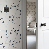 Bronze Light Switch On Blue And White Floral Wallpaper With White Door Ajar And White Chair In The Distance