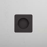 Black square wall fixture with bronze-edged circular center, mounted on a textured white wall.