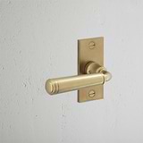 Digby Short Plate Fixed Door Handle in Antique Brass with a sleek rectangular backplate on a textured white wall.
