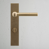 Harper Long Plate Sprung Door Handle & Thumbturn in antique brass on a textured white wall.