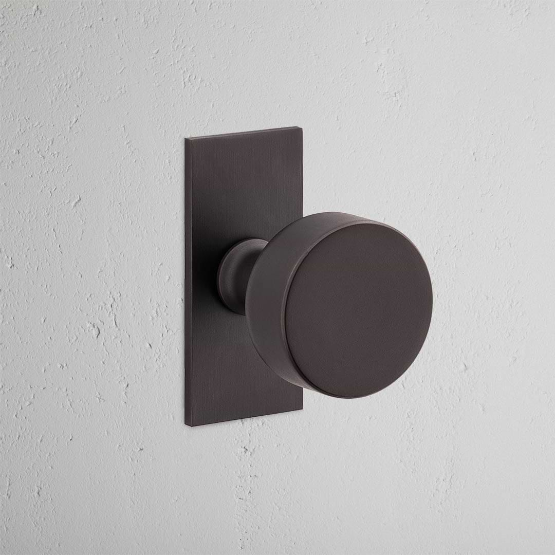 Bronze Onslow Short Plate Fixed Door Knob on a textured white wall.