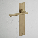 Clayton Long Plate Sprung Door Handle in antique brass with elegant rectangular backplate on a white wall.