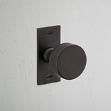 Bronze Onslow Short Plate Sprung Door Knob on rectangular plate against a textured white wall.
