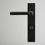 Harper Long Plate Sprung Door Handle & Thumbturn in bronze against a textured white wall, showcasing elegant design.