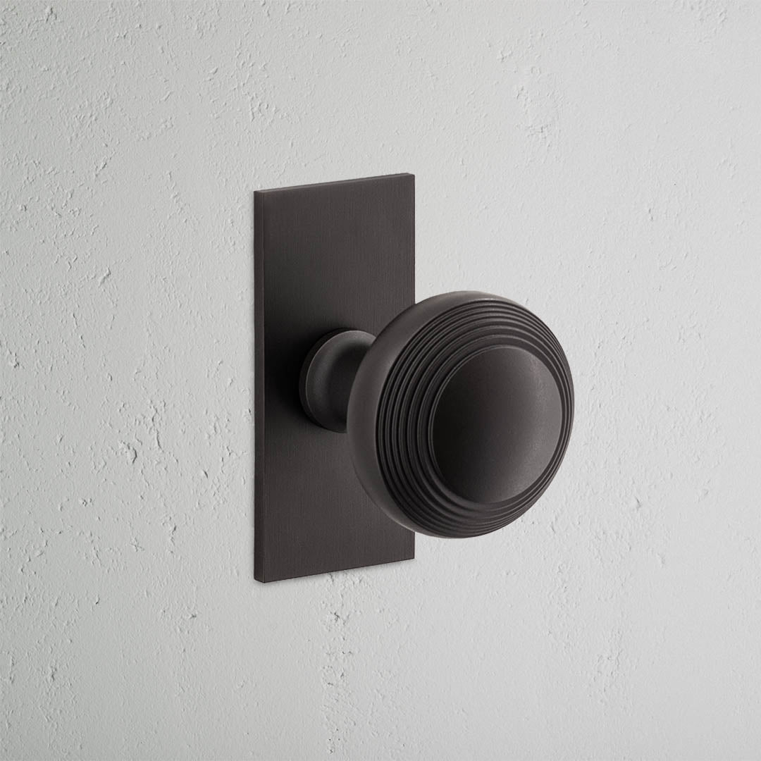 Bronze Poplar Short Plate Door Knob with circular grooves, fixed on a white wall.