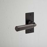 Digby Short Plate Sprung Door Handle in Bronze displayed on a white wall, offering a touch of elegance.