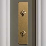 Antique Brass 2G Architrave Two Way Toggle Switch in a classic design, shown on a gray wall.