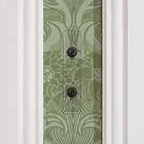 Clear bronze 2G architrave two-way toggle switch against ornate green floral wallpaper with a white border.