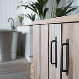 Bronze Kilburn Furniture Handle 128mm on a cabinet with bathroom and plants in the background.