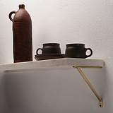 Antique Brass Brixton Shelf Bracket 150mm supporting a white marble stone shelf fixed onto a textured white wall with dark coloured pottery placed on top.