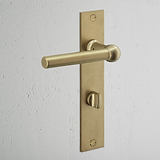 Harper Long Plate sprung door handle & thumbturn in antique brass on a white textured wall.