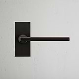 Bronze Clayton Short Plate Sprung Door Handle mounted on a textured white wall.