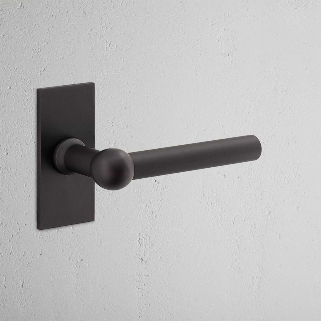 Harper Short Plate Fixed Door Handle in bronze against a white textured wall, offering a sleek and modern touch.