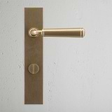 Antique Brass door handle and thumbturn with a rectangular backplate on a light gray textured wall.