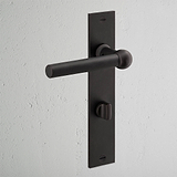 Harper Long Plate Sprung Door Handle & Thumbturn in bronze on a white textured wall.