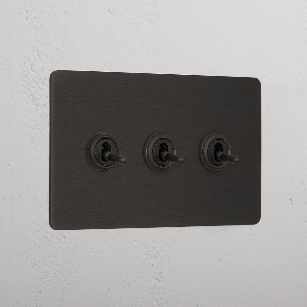 Bronze panel with three retractive toggle switches (3G) mounted on a white wall.