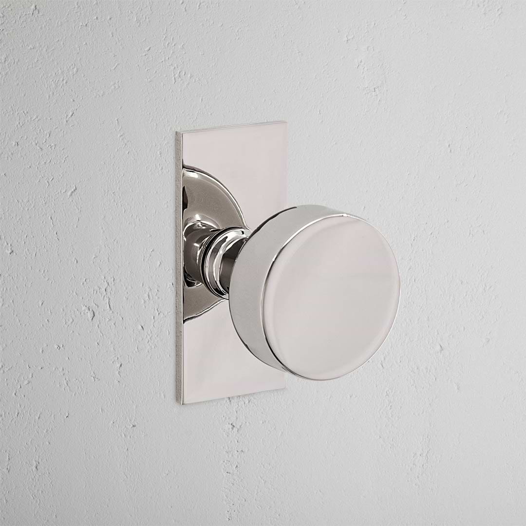 Polished Nickel 'Onslow Short Plate Fixed Door Knob' on a sleek rectangular plate against a light gray wall.
