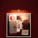 Abstract painting with geometric shapes lit by a Taylor Picture Light on a red wall.