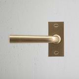 Apsley Short Plate Sprung Door Handle in antique brass, mounted on a textured white wall.