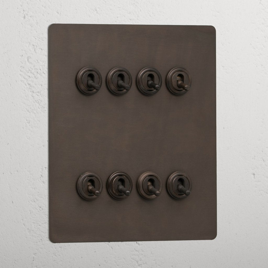 Bronze toggle light switch panel with 8G retractive switches mounted on a textured wall.